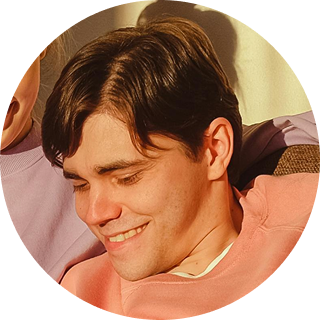 Young man with dark hair smiling, wearing a coral-colored shirt against a warm-toned background