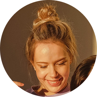 Woman with blonde hair in a top bun, smiling warmly with eyes closed against a brown background